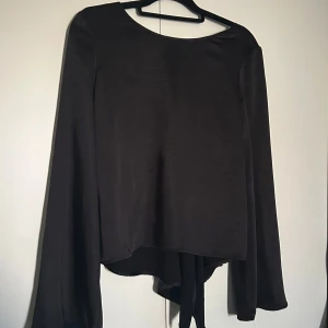 Svart blus från Gina tricot - Snygg svart omlottopp med långa, vida ärmar och knytband framtill. Toppen har en lätt glansig finish och en djupare urringning i ryggen. Perfekt för dig som gillar en trendig och lite dramatisk look. Använd 1 gång