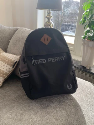 Svart ryggsäck från Fred Perry - Snygg svart ryggsäck från Fred Perry med klassisk logga och reflekterande detaljer på sidorna. Tillverkad i slitstark textil med dubbla dragkedjor och en brun läderpatch upptill. Perfekt för skolan eller stan med sin stilrena design och praktiska fack.