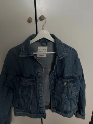 Blå jeansjacka från Pull&Bear - Snygg blå jeansjacka från Pull&Bear med klassisk krage, två bröstfickor med lock och knappar samt knappstängning framtill. Jackan har en avslappnad passform och tidlös design som passar till det mesta. Perfekt för dig som gillar streetstyle och denim.