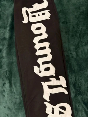 Svarta YoungLA mjukis/gymbyxor - Säljer ett par svarta mjukis/gym byxor från YoungLA med stor vit text längs benet. Byxorna har resår i midjan och en bakficka med en liten YoungLA-label. Perfekta för en chill och street-inspirerad look i eller utanför gymmet.