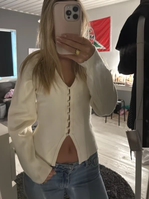 Vit cardigan med knappar - Trendig vit cardigan med djup V-ringning och dekorativa knappar framtill. Modellen har långa ärmar och en lätt utsvängd nederkant som ger en snygg siluett. Perfekt att styla med jeans för en avslappnad look.