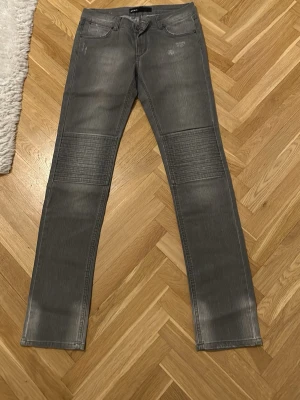 Grå bikerjeans med ribbade knän - Säljer ett par grå bikerjeans med ribbade detaljer över knäna och diskreta slitningar. Jeansen har fem fickor och raka ben. Perfekta för en cool och avslappnad stil. Materialet är troligtvis bomull med stretch för extra komfort.