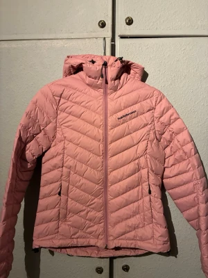 Ljusrosa dunjacka från Peak Performance - Snygg ljusrosa dunjacka från Peak Performance med huva och dragkedja framtill. Jackan har två sidofickor med dragkedja och ett quiltat mönster i chevron-stil. Perfekt för våren och riktigt lätt att bära. Inga defekter eller hål. 
