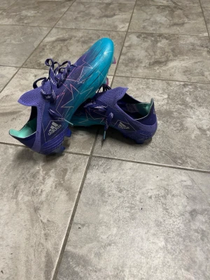 Adidas fotbollsskor - Snygga Adidas fotbollsskor i blått och lila med turkosa detaljer och grafiskt mönster i vitt och rosa. Skorna har snörning och låg profil, samt slitstark syntetisk ovandel som ger bra bollkontroll. Perfekta för dig som vill sticka ut på planen. Det är elite modellen och dem är i väldigt bra skick! Priset går att diskutera!