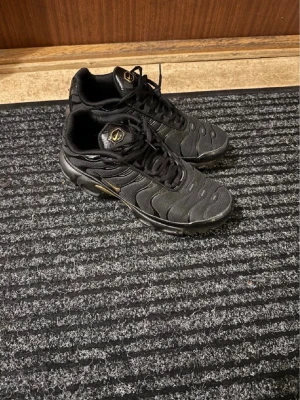 Svarta Nike sneakers med gulddetaljer - Snygga svarta Nike sneakers med gulddetaljer och klassisk snörning. Skorna har en sportig design med rund tå och platt sula. Materialet är syntet och textil, vilket ger en lätt och bekväm känsla. Perfekta för dig som gillar streetwear och stilrena färger. Det finns hål 