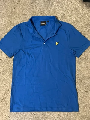Blå pikétröja från Lyle & Scott - Blå pikétröja från Lyle & Scott med klassisk krage, korta ärmar och knappslå. Tröjan har det ikoniska gula örnemblemet broderat på bröstet och är gjord i mjuk bomull för en bekväm känsla.