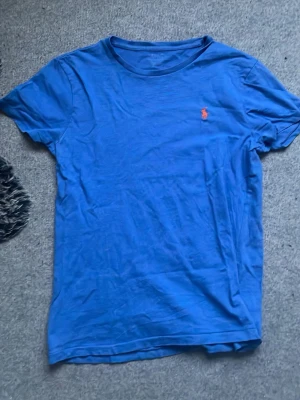 Blå t-shirt från Ralph Lauren - Snygg blå t-shirt från Ralph Lauren med klassisk passform och korta ärmar. T-shirten har det ikoniska röda Polo-logotypen broderad på bröstet. Tillverkad i mjuk bomull som känns skön mot huden. Perfekt till jeans eller shorts.