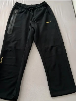 Svarta NOCTA x Nike mjukisbyxor - Säljer ett par svarta mjukisbyxor från Nike och NOCTA-kollektionen. Byxorna har gul logga, dragsko i midjan och en unik ficka med dragkedja på sidan. Broderade detaljer i gult på benen och relaxed passform. Perfekta för chill eller streetwear.