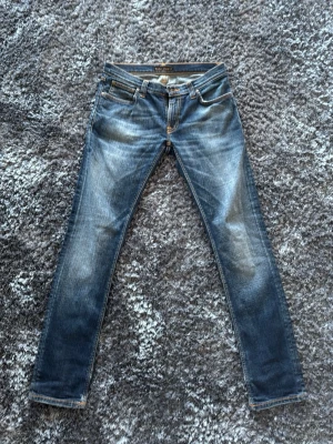 Blå slim jeans från Nudie Jeans - Säljer ett par blå slim fit jeans från Nudie Jeans med klassisk femficksdesign och snygga slitningar på framsidan. Jeansen har en ganska slimfit passform och är tillverkade i denim med kontrastsömmar och läderpatch bak. Perfekta för dig som gillar en modern och stilren look. OBS STORLEK - W29 L32