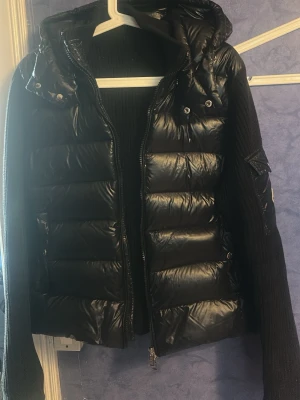 Svart dunjacka från Moncler - Snygg svart dunjacka från Moncler med glansig finish, stickade ärmar och huva. Jackan har dragkedja framtill, fickor med lock och Moncler-logga på ena ärmen. Perfekt för kalla vinterdagar och riktigt trendig look.