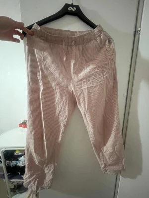 Randiga ljusrosa loungewear-byxor - Mysiga loungewear-byxor i ljusrosa med tunna vita ränder. Byxorna har resår i midjan med dragsko för extra komfort och raka ben. Perfekta för chill hemma eller när du vill vara bekväm. Materialet känns lätt och luftigt.