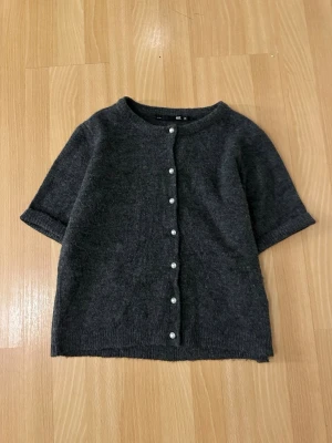 Mörkgrå kofta med pärlknappar - En stilren mörkgrå kofta från GU med korta ärmar och rund hals. Koftan har pärlknappar framtill och är tillverkad i ett mjukt stickat material som ger en skön känsla. Perfekt för att skapa en enkel och snygg look.