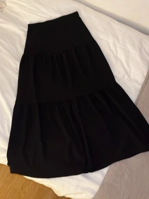 Svart lång kjol i mjukt tyg - En svart lång kjol med volangdetaljer och bred linning. Kjolen är sydd i ett mjukt, stretchigt material som ger en avslappnad och skön känsla. Passar perfekt till både sneakers och boots för en trendig look.