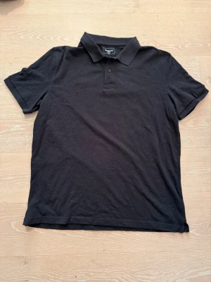 Svart pikétröja från Dressmann - Klassisk svart pikétröja från Dressmann med krage och knappar framtill. Tröjan har korta ärmar och är tillverkad i bomull, perfekt för en clean och enkel stil.