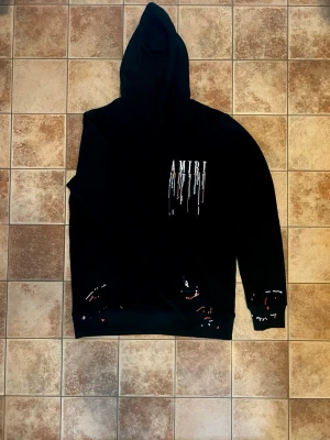 Svart hoodie från Amiri med färgstänk - Säljer en svart hoodie från Amiri med färgstänk och droppdetaljer i flera färger på bröstet och runt muddar. Hoodien har huva och stor magficka framtill. Perfekt för dig som gillar streetwear och vill sticka ut med en unik design.