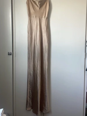 Champagnefärgad långklänning i satin - Elegant långklänning i champagnefärgad satin med smala axelband och figurnära passform. Klänningen har en enkel, stilren design utan synliga detaljer och faller mjukt längs kroppen. Perfekt för dig som vill ha en minimalistisk och lyxig look. Använd 1 gång! Förutom fläck visad på bilden är den i bra skick!