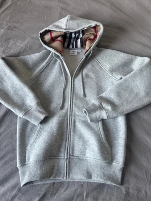 Grå zip hoodie från Burberry - Snygg grå zip hoodie från Burberry med klassiskt rutigt foder i huvan och broderad logga på bröstet. Tillverkad i mjuk bomullsblandning, har dragsko i huvan och två fickor framtill. Perfekt för en avslappnad och stilren look.