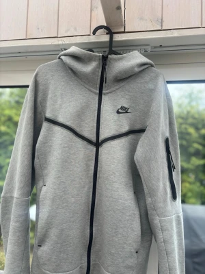 Grå Nike Tech Fleece hoodie🩶 - Snygg grå Nike Tech Fleece hoodie med svart dragkedja och svarta detaljer. Hoodien har huva, fickor på sidorna och en extra ficka med dragkedja på ärmen. Tillverkad i mjukt och stretchigt material som är perfekt för chill eller träning. Nike-logga på bröstet.