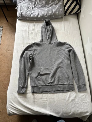 Grå basic hoodie med ficka - En enkel och stilren grå hoodie med huva och stor magficka. Hoodien har ribbade muddar vid ärmslut och nederkant. Perfekt för chill dagar eller när du vill ha en clean look. Materialet känns mjukt och bekvämt, troligen bomullsblandning.