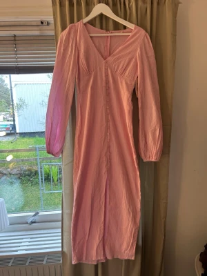 Ljusrosa långklänning med knappar - Söt ljusrosa långklänning från NA-KD med v-ringning och långa, lätt puffade ärmar. Klänningen har dekorativa knappar framtill och är tillverkad i ett tunt, lätt strukturerat material. Perfekt för vår och sommar.
