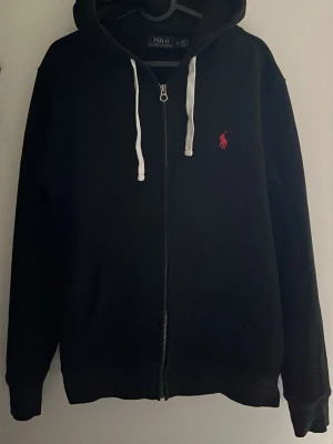 Svart hoodie från Polo Ralph Lauren - Svart hoodie med dragkedja från Polo Ralph Lauren. Klassisk design med vit dragsko i huvan och röd broderad logga på bröstet. Mjuk bomullskänsla och ribbade muddar. Perfekt för en chill och stilren look.