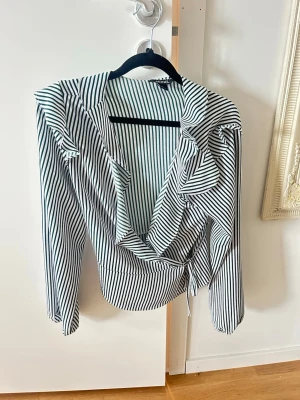 Randig omlottblus från Monki - Snygg omlottblus från Monki med vita och mörkgröna smala ränder. Blusen har långa ärmar, volangdetaljer vid axlar och omlottknäppning i midjan. Perfekt för en trendig och stilren look. 