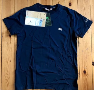 Mörkblå Burberry t-shirt i bomull - Snygg mörkblå t-shirt från Burberry med klassisk passform. T-shirten har korta ärmar, rund halsringning och en liten broderad Burberry-logga på bröstet. Tillverkad i mjuk bomull och perfekt för en stilren vardagslook.