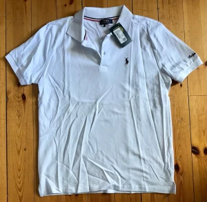 Vit pikétröja från Ralph Lauren - Klassisk vit pikétröja från Ralph Lauren med broderad logga på bröstet och diskreta ränder i kragen. Tröjan har korta ärmar och knappar vid halsen. Perfekt för en stilren och sportig look.