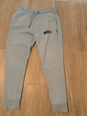 Ljusblå mjukisdress från Hoodrich - Snygg ljusblå mjukisdress från Hoodrich med broderad logga i lila och rosa nyanser på låret. Byxorna har dragsko i midjan, muddar vid bensluten och två sidofickor. Perfekta för en chill och avslappnad stil. Skriv privat för mer info!
