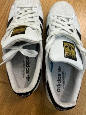 Adidas Superstar vita med svarta ränder - Klassiska Adidas Superstar sneakers i vitt läder med svarta ränder och guldiga detaljer på plösen. Skorna har den ikoniska snäckformade tån och Adidas logga på sidan och plösen. Perfekta för en streetwear-look.