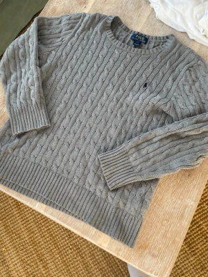Grå kabelstickad tröja Polo Ralph Lauren - English: Classic grey cable-knit sweater from Polo Ralph Lauren. Features a soft cotton knit, crew neckline, ribbed cuffs and hem, and the iconic embroidered logo on the chest. A timeless and versatile piece, perfect for both casual and smart outfits. Fits approximately ages 12–14, men’s XS, or around 160–165 cm depending on desired fit. Comfortable and ideal for layering.