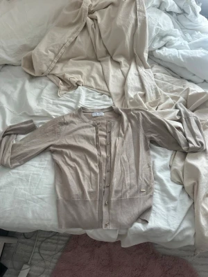 Beige kofta från Holly & Whyte - Snygg beige kofta från Holly & Whyte med knappar framtill. Lätt och tunn i materialet, perfekt att slänga över en t-shirt. Klassisk och enkel design som passar till det mesta.