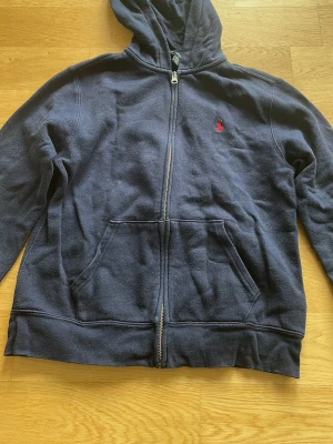 Mörkblå hoodie från Ralph Lauren - Mörkblå hoodie med dragkedja från Ralph Lauren. Klassisk design med huva, kängurufickor och röd broderad logga på bröstet. Ganska nopprig 