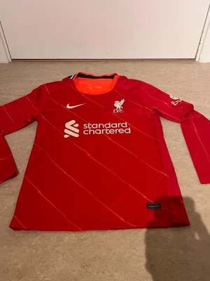 Liverpool långärmad matchtröja Nike - Röd långärmad Liverpool FC matchtröja från Nike med vita detaljer och klubbmärke på bröstet. Tröjan har tryckt sponsorlogga och tunna diagonala linjer i designen. Perfekt för fotbollsträning eller att bära på läktaren.