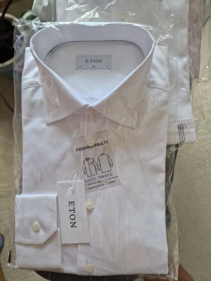 Vit skjorta från Eton slim fit - Klassisk vit skjorta från Eton i slim fit-modell. Skjortan har knappar framtill, lång ärm och stilren krage. Tillverkad i bomull för en fräsch och bekväm känsla. Perfekt för dig som gillar en clean och tidlös look.