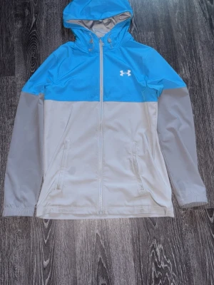 Blå och grå vindjacka från Under Armour - Snygg tvåfärgad vindjacka från Under Armour i blått och ljusgrått. Jackan har huva med dragsko, hel dragkedja framtill och två sidofickor. Perfekt för blåsiga dagar och sportiga outfits.