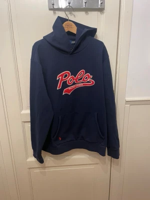 Marinblå hoodie från Polo Ralph Lauren - Snygg marinblå hoodie från Polo Ralph Lauren med stor röd och vit broderad logga på bröstet. Klassisk känguruficka framtill och huva med dragsko. Perfekt för en avslappnad och sportig stil.
