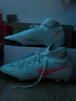 Nike phantom elite fotbollsskor - Säljer ett par turkosa Nike phantom elite fotbollsskor med hög krage och röd swoosh på sidorna. Skorna har snörning framtill och är designade för bra grepp på planen. Tillverkade i syntetmaterial och har en modern, strömlinjeformad look.