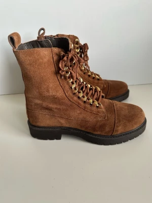 Bruna mockakängor med gulddetaljer, storlek 36 - Snygga bruna kängor i 100% äkta mocka från Copenhagen Shoes i storlek 36. Tidlös modell med grov sula och guldfärgade öljetter. Kängorna har snörning framtill, dragkedja på sidan och en praktisk ögla bak. Lika snygga till klänning på sommaren som till byxor på vintern. Härliga kvalitetsskor som kommer hålla säsong efter säsong. Nypris 1795:-. Mycket gott skick, som nya. 