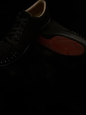 Svarta sneakers med nitar och röd sula - Säljer ett par svarta sneakers från Christian Louboutin med coola svarta nitar på tån och klassisk röd sula. Skorna har svarta skosnören och en låg profil. Insidan är ljusbeige och materialet är troligtvis läder eller mocka.