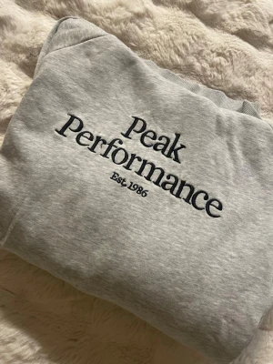 Grå hoodie från Peak Performance - Grå hoodie från Peak Performance med svart broderad logga och text 'Est. 1986' på bröstet. Klassisk modell med mjukt material och ribbade muddar vid ärmslut och nederkant. Perfekt för en avslappnad och sportig stil.