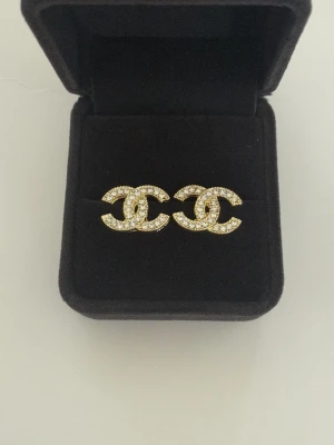 Guldiga CC örhängen med strass - Snygga CC örhängen i guldton med klassiska CC-loggan täckt av glittrande strass. Perfekt accessoar för att lyfta din stil med en lyxig touch. Kommer i svart ask.
