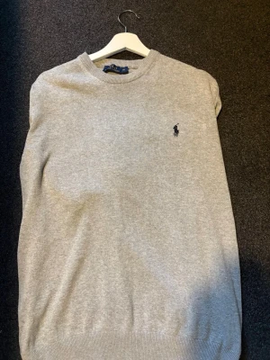 Grå stickad tröja från Polo Ralph Lauren - Snygg grå stickad tröja från Polo Ralph Lauren med rund halsringning och klassisk broderad logga på bröstet. Tröjan har ribbade muddar vid ärmslut och nederkant, perfekt för en stilren look. Materialet är mjukt och bekvämt.