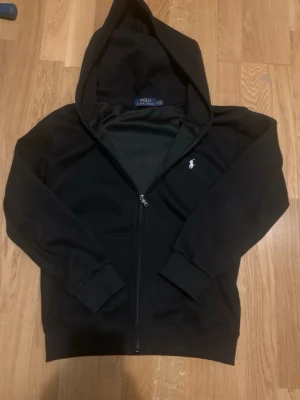 Svart zip up Polo Ralph Lauren tröja - Svart hoodie med dragkedja från Polo Ralph Lauren. Klassisk design med huva, ribbade muddar och vit broderad logga på bröstet. Perfekt för en chill och stilren look. Materialet är mjukt och bekvämt, passar till det mesta i garderoben.