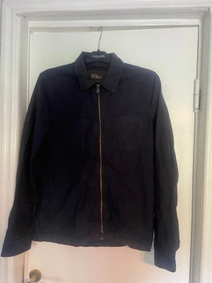 Svart overshirt från oscar jacobson - Stilren svart overshirt från oscar jacobson med klassisk krage och dragkedja framtill. Overshirten har två bröstfickor och raka ärmar med knapp vid ärmslut. Perfekt att slänga över en t-shirt för en avslappnad look.