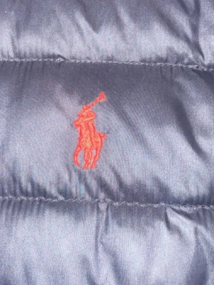 Mörkblå dunväst från Polo Ralph Lauren - Snygg svart dunväst från Polo Ralph Lauren med klassisk röd broderad logga på bröstet. Västen har en quiltad design och är tillverkad i glansigt material som ger en exklusiv känsla. Perfekt för lager-på-lager och streetwear-stil.