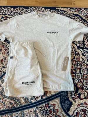 Essentials Fear of God set grå - Säljer ett grått set från Essentials Fear of God med t-shirt och shorts. Båda plaggen har svart logotyptryck framtill. T-shirten har rund hals och shortsen har resår i midjan med dragsko. Mjukt och bekvämt material, perfekt för en avslappnad stil.