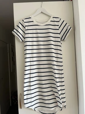 Randig t-shirtklänning från JDY - Vit t-shirtklänning från JDY med marinblå horisontella ränder. Klänningen har rund halsringning och korta ärmar, tillverkad i mjuk bomull. Perfekt för en avslappnad och stilren look.