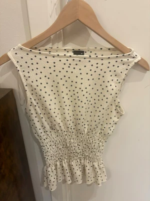 Prickig ärmlös topp från Gina Tricot - Supersöt ärmlös topp från Gina Tricot i vitt med svarta prickar. Toppen har smockad midja och volangkant nedtill! Använd endast fåtal gånger💕