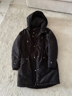 Svart parkas från Vero Moda - Svart parkas från Vero Moda med huva, dragkedja och tryckknappar framtill. Jackan har flera fickor med lock och dragkedja samt justerbar midja. Perfekt för kalla dagar med sin vadderade insida och klassiska design.